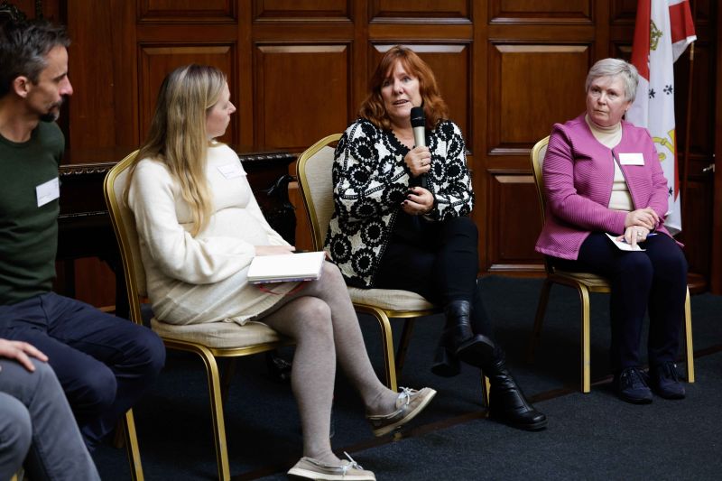 Four participants seated, with one participant speaking into a microphone and the other three listening.